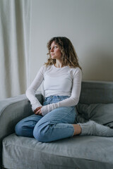 Obraz premium Pensive young woman wearing casual clothes sitting on a comfortable sofa at home, looking away from the camera with a thoughtful expression