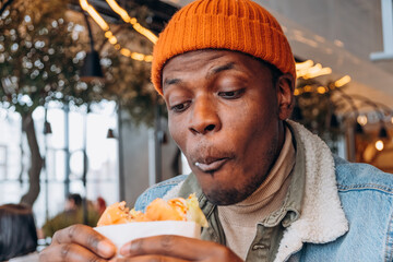 Concentrated dark-skinned man wearing orange beanie blowing air while eating burger in fast food...