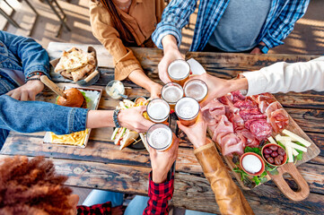 Friends cheering beer glasses on wooden table covered with food at brewery pub garden - Top view of...