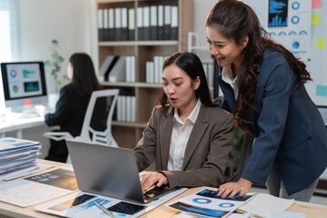 Two asian businesswomen working together, analyzing charts and data on a laptop in a modern office environment, collaborating on a project