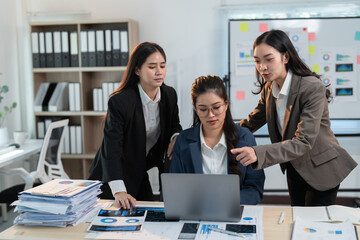 Three asian businesswomen are working together in the office, analyzing charts and graphs on a laptop and printed documents, collaborating on a project