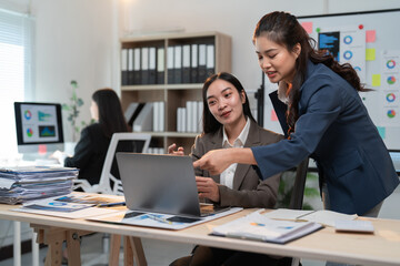 Obraz premium Two asian businesswomen are discussing work and analyzing financial graphs using a laptop in a modern office, showcasing teamwork and collaboration
