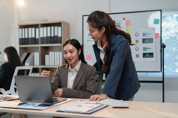 Obraz premium Two asian businesswomen are discussing work using a laptop in a modern office, collaborating on a project and analyzing data on the computer screen