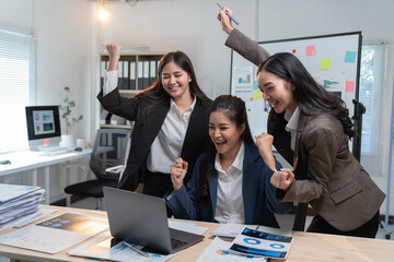 Three enthusiastic businesswomen are raising their arms in celebration. Expressing joy and excitement over positive results achieved while working together on a laptop in a modern office environment