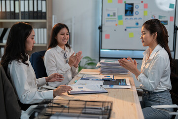 Three asian businesswomen are sitting at a desk in a modern office, clapping their hands after a successful presentation, celebrating their achievements and teamwork