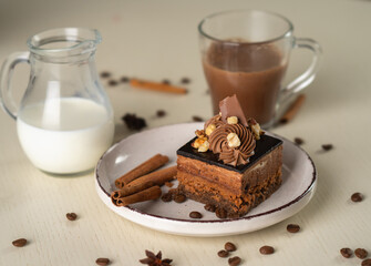 A piece of chocolate dessert with nuts and cream, served on a plate with cinnamon sticks, next to a milk jug and a cup of cocoa