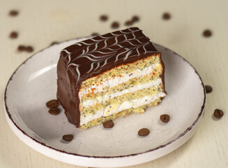 A slice of sponge cake with poppy seeds, cream filling, and chocolate glaze on a white plate with coffee beans