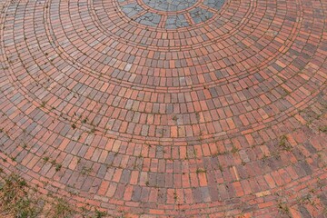 Image shows a circular brick pavement or flooring, with vegetation growing through some joints The...