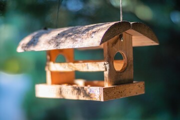 A weathered birdhouse hangs on a hook or branch, with a sloping roof and square base It has an upper front opening for birds The setting appears to be outdoors, possibly in daylight