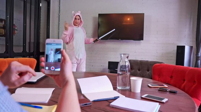 Man in pink onesies plastic toy sword speaks at corporate meeting, conference, presents creative advertising ideas, hand holds smartphone, film eccentricities of freak on camera incriminating evidence
