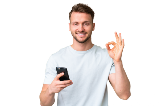 Young handsome caucasian man over isolated chroma key background using mobile phone and doing OK sign