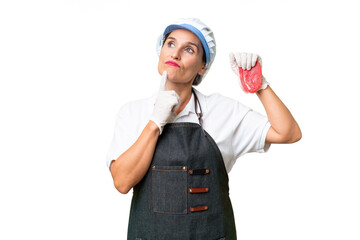 Butcher woman wearing an apron and serving fresh cut meat over isolated background having doubts while looking up