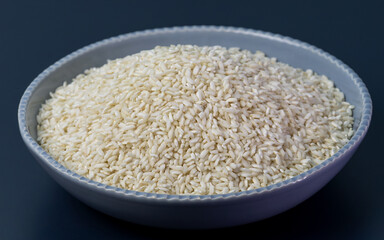 Uncooked Carnaroli risotto rice in blue bowl on a dark blue table. 