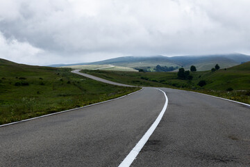 A winding road stretches through a hilly landscape with green fields and scattered trees under an overcast sky