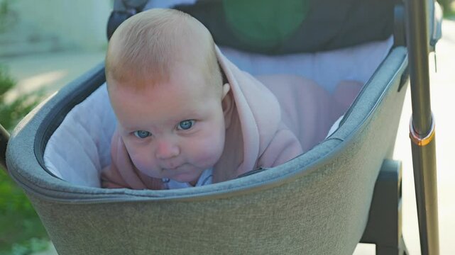 A baby with bright blue eyes in a pink sweatshirt smiles mischievously while lying on his stomach in a grey pram. Close-up portrait, outdoors with soft natural light