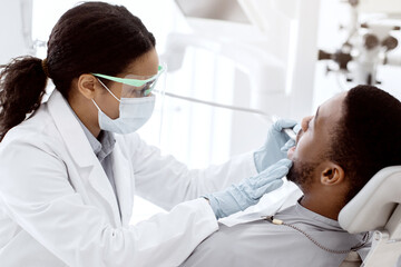 Portrait Of Black Male Patient Getting Teeth Treatment With Dentist At Modern Clinic, Sitting In Chair In White Cabinet. Professional Stomatologist Lady Using Dental Drill Tool For Young Man