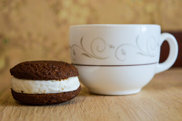 white cup on the surface sits next to a chocolate cookie sandwich filled with white cream food and beverage dessert products recipe cup of tea or coffee and a treat elegant tableware artisan cookies