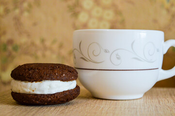 white cup on the surface sits next to a chocolate cookie sandwich filled with white cream food and beverage dessert products recipe cup of tea or coffee and a treat elegant tableware artisan cookies