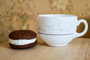 white cup on the surface sits next to a chocolate cookie sandwich filled with white cream food and beverage dessert products recipe cup of tea or coffee and a treat elegant tableware artisan cookies