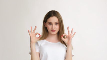 Helpful girl. Calming sign. Enthusiastic teenager in white t-shirt showing okay approval gesture isolated on light empty space background.