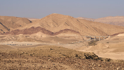 Bridge above a road leading inside the Big Crater in Negev Desert. Chain of varicolored mountain ridges on the background. Dramatic  colorful desert landscape. Cars and tents in Ein Yorkeam campground