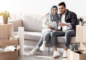 Happy muslim family sitting on couch in new apartment with digital tablet among boxes with...