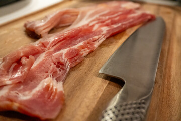 Close up of Raw Slices of Bacon on a Wooden Cutting Board with a Stainless Steel Knife