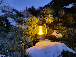 A glowing small decorative lantern stands in the snow on the branches of a fir tree. The concept of New Year and Christmas. © Sofya