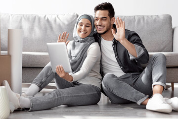 Obraz premium Happy young loving middle-eastern family husband and wife in hijab having online party with friends, showing their new apartment, sitting on floor among moving boxes, using digital tablet