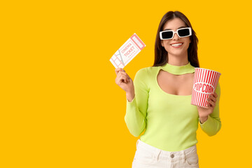 Young happy woman in 3D cinema eyeglasses with popcorn bucket and ticket on yellow background
