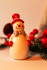 Holiday New Year's decoration, decor of a ceramic snowman with a red hat and a bow on a background of Christmas tree branches, with red berries and cones.