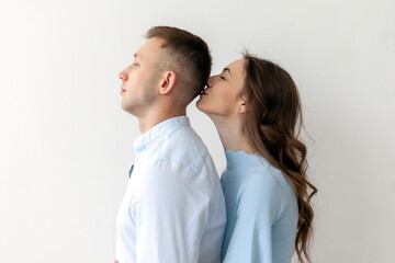 young couple in love stands and hugs against the background of white isolated wall, woman kisses man on the back of the head, copy space