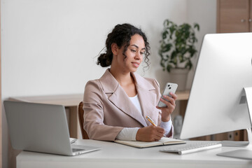 Young African-American QA engineer with mobile phone and notebook at her workspace in office