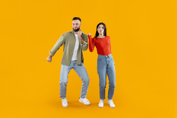 A young couple dances playfully against a bright yellow backdrop. They express joy and connection through their movements, showcasing a lighthearted moment together while dressed casually.