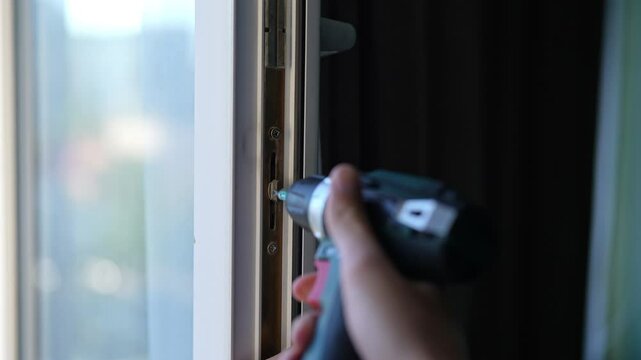 Close-up of professional repairman worker tightening screws on window lock mechanism using cordless screwdriver during maintenance service, slow motion. Concept of plastic window maintenance.