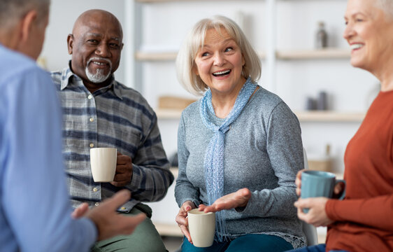 Multiracial group of positive senior men and women sitting in circle, drinking tea and having conversation, smiling and laughing, having home party or enjoying time at nursing home, closeup