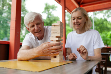 elderly couple of seniors in white t-shirts playing board game in park outdoors, old man and woman building a tower of wooden bricks and competing in a game of balance