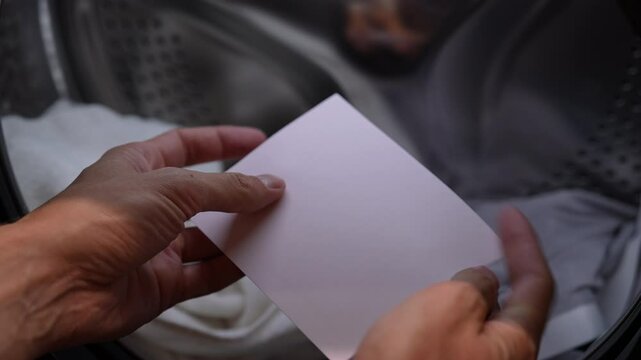 Male housekeeper using eco-friendly pink laundry detergent sheet to do laundry in open washing machine, preparing to do laundry with innovative cleaning products, closeup, slow motion.