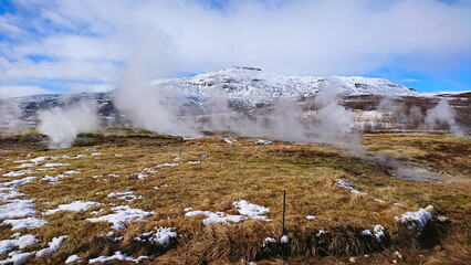 Haukadalur, Iceland - 3.24.2018: Geysers with steam coming out constantly in between a grassland with snow and dried meadows under a cloudy blue sky with a snow mountain at the back before pandemic