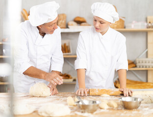 Experienced aged baker guiding eager young female apprentice, kneading and rolling dough together in small artisan bakery filled with freshly baked bread..