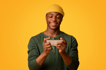 Mobile Gaming Concept. Excited Young Black Guy Playing Online Games On Smartphone, Cheerful African American Man Having Fun With Mobile Phone While Standing Over Yellow Studio Background, Free Space