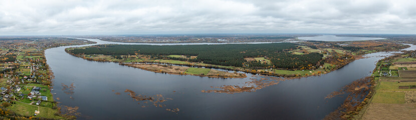 Wide aerial view of a river curving through a landscape with forests, fields, and residential areas. Overcast sky adds a muted tone to the scene.