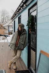 stylish young woman in warm winter clothes against the background of a small wooden house decorated with Christmas decorations