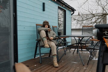 stylish girl in a warm jacket and hat in the yard of a tourist house is grilling meat and drinking coffee, enjoying a rural vacation and natural scenery