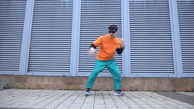 young crazy guy dances hip hop on city street and moves to the music against the background of wall, male dancer performs freestyle in an orange t-shirt outdoors