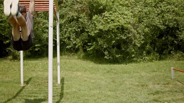 A young man performs leg raises at an outdoor gym. He hangs from the monkey bars and lifts his legs up, holding them straight out in front of him.