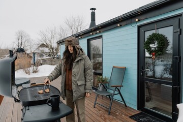 Fototapeta premium winter vacation in the backyard of a country tourist house. A young stylish woman is grilling meat on a gas grill for her friends and family