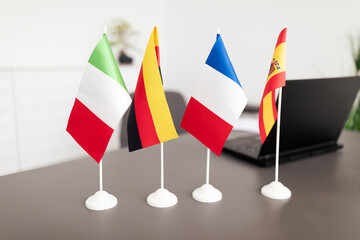 Four small national flags, including Italy, Germany, France, and Spain, are arranged on a desk in a contemporary office. The workspace features a laptop and a clean design, reflecting professionalism.