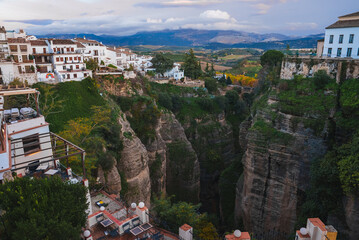 View of Ronda, Spain, featuring El Tajo Gorge, whitewashed buildings on cliffs, lush greenery, rugged rocks, rolling hills, and a partly cloudy sky.