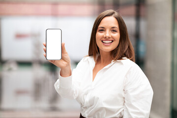 A young businesswoman smiles confidently while holding up her smartphone in a contemporary office setting. The background reflects a vibrant corporate atmosphere focused on work.